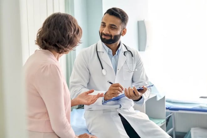 Male doctor sits in bed smiling at patient while writing on clipboard