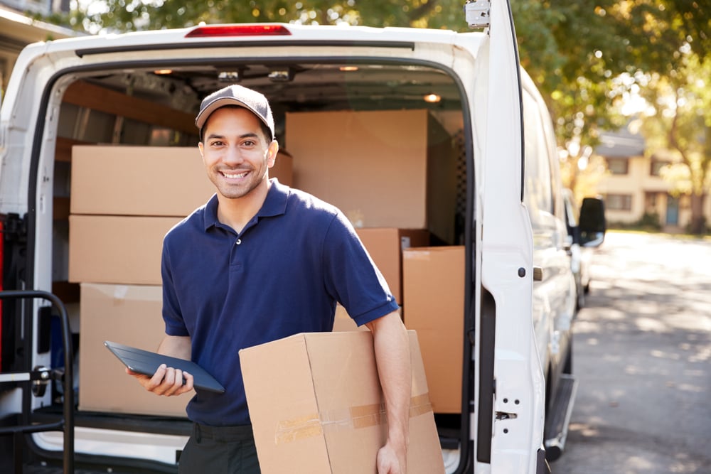 Delivery driver holding brown box and clipboard smiles at camera